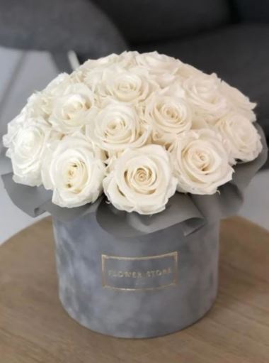 A luxurious arrangement of pristine white roses tightly packed in a round, grey velvet hatbox with a gold-embossed label.