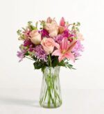 A lush floral arrangement in a clear glass vase featuring pink roses, pink lilies, and purple chrysanthemums against a plain white background.