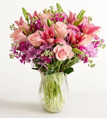 A lush and vibrant floral arrangement in a clear glass vase, featuring pink roses, pink lilies, and purple chrysanthemums with green foliage against a white background.