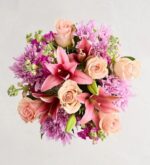 A top-down view of a vibrant floral bouquet featuring a mix of pink lilies, peach roses, and purple chrysanthemums arranged in a dense, circular composition.
