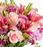 A lush and vibrant floral bouquet featuring a mix of light pink roses, deep pink lilies, and purple chrysanthemums with green foliage against a white background.