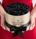 A person in a red dress holding a round beige hatbox tied with a black satin ribbon, filled with preserved black roses. The box is labeled "MAGNIFICENT ROSES".