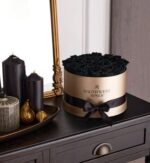 Round gold hat box with black ribbon labeled “Magnificent Roses” filled with black preserved roses, styled on a dark wood dresser next to black pillar candles and gold decor accents.