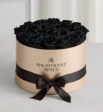 Luxury round gold gift box filled with preserved black roses, wrapped with a black satin ribbon and bow. The box features "Magnificent Roses" branding on the front.