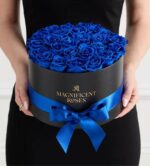 A person in a black dress holds a round black hat box filled with vibrant blue velvet roses. The box has gold "Magnificent Roses" lettering and is wrapped with a wide royal blue satin ribbon tied in a bow.