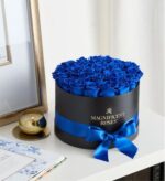 A round black hatbox tied with a vibrant blue satin ribbon, filled with preserved royal blue velvet roses, displayed on a white shelf next to a gold candle and framed artwork