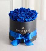 Luxury arrangement of vibrant blue velvet preserved roses densely packed in a round black gift box with gold “Magnificent Roses” logo, wrapped with a glossy royal blue satin ribbon and bow, displayed on a white marble surface.
