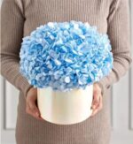 Person in a beige ribbed sweater holding a round white hatbox filled with a lush bouquet of light blue hydrangea blooms.