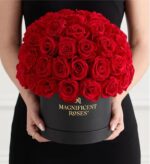 Woman in black dress holding a round black box filled with vibrant red roses arranged in a dome shape, with gold “MAGNIFICENT ROSES” logo on the box.