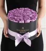 Person holding a round black gift box filled with fresh lavender roses, wrapped with a satin lavender ribbon and bow, gold "Magnificent Roses" logo on the front
