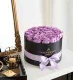 A round black gift box filled with fresh lavender roses, wrapped with a light purple satin ribbon and gold “Magnificent Roses” logo. The box sits on a white surface next to a gold-framed mirror and a black tray holding a gold candle holder and gift tags.