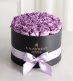 Round black gift box filled with fresh lavender roses, wrapped with a pale lilac satin ribbon and bow, with "Magnificent Roses" gold logo on the front, displayed on a white surface with soft white curtains in the background.