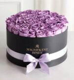 Round black gift box filled with dozens of fresh lavender roses, wrapped with a pale lilac satin ribbon and bow. Gold “MAGNIFICENT ROSES” logo printed on the front of the box.