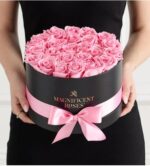 A person wearing a black dress holds a round black gift box filled with tightly arranged pink roses, wrapped with a light pink satin ribbon and bow. The box features gold “MAGNIFICENT ROSES” branding.