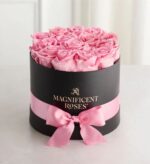Round black hat box filled with fresh pink roses, wrapped with a pink satin ribbon and bow, with gold “Magnificent Roses” logo on the front.