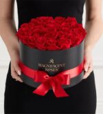 A person in a black dress holding a round black box filled with vibrant red roses, tied with a red satin ribbon. The box is labeled “Magnificent Roses” in gold lettering.
