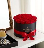 A round black hat box filled with vibrant red roses, wrapped with a red satin ribbon and bow. The box reads “Magnificent Roses” in gold lettering. It sits on a white vanity next to a lit candle and gold-framed mirror.