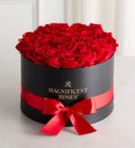 Luxury round black gift box filled with fresh red roses, wrapped with a glossy red satin ribbon and bow. Gold "Magnificent Roses" logo on front.