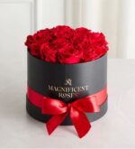 A round black gift box with a glossy red satin ribbon and bow, filled with a dense arrangement of vibrant red roses. The box is embossed with "MAGNIFICENT ROSES" in gold lettering and sits on a light marble surface with soft white curtains in the background.