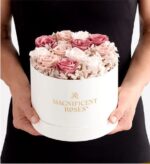 Person in black dress holding a round white box with gold "Magnificent Roses" logo, filled with preserved roses in blush pink, mauve, and ivory tones, accented with hydrangea.
