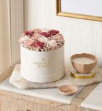 Round white hat box with gold "Magnificent Roses" logo, filled with preserved roses in blush, dusty rose, and ivory tones mixed with hydrangeas. The box sits on a marble tray next to a taupe ceramic candle and small dish on a light wood table with white paneled wall.