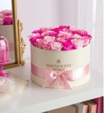 Round cream-colored hat box filled with a dense arrangement of light pink and hot pink roses, tied with a pale pink satin ribbon and bow. The box has “Magnificent Roses” printed in gold on the front. It sits on a white shelf next to a gold-framed mirror and a pink glass candle holder.