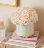 A dome of preserved blush roses arranged in an iridescent white ceramic vase, styled on a stack of books beside a lit candle and gold-framed wall art.