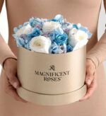 Hands holding a round beige gift box labeled “Magnificent Roses” filled with preserved white, sky blue, and lavender roses with light blue hydrangea accents.