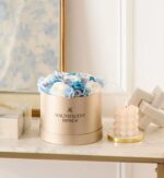 Round gold box of preserved white and light blue roses on a marble surface, styled with a lit bubble candle and decorative accents