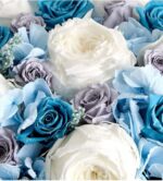 Close-up of a preserved floral arrangement featuring large white garden roses, deep teal and dusty lavender roses, pale blue hydrangeas, and small white filler flowers, arranged in a soft coastal color palette.