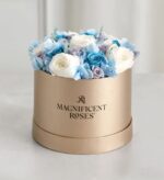 Round gold hat box with “Magnificent Roses” logo, filled with preserved white, light blue, and lavender roses and hydrangeas, arranged on a light marble surface.