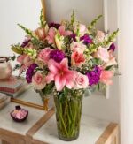 A lush floral arrangement in a glass vase featuring pink lilies, light pink roses, purple stock flowers, and green snapdragons, displayed on a marble-top console table.