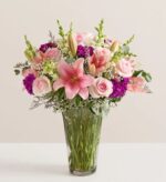 A lush floral arrangement in a glass vase featuring pink lilies, light pink roses, purple stock flowers, and white waxflowers with green foliage.