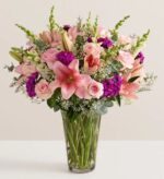A lush and premium floral bouquet featuring large pink lilies, light pink roses, and vibrant purple stock flowers, arranged with white baby's breath and green eucalyptus in a clear glass vase.