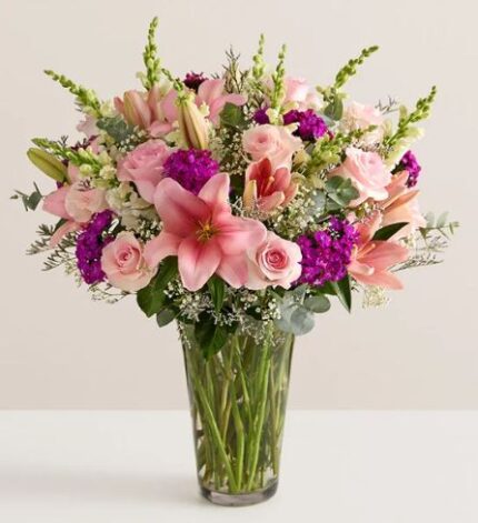 A lush and premium floral bouquet featuring large pink lilies, light pink roses, and vibrant purple stock flowers, arranged with white baby's breath and green eucalyptus in a clear glass vase.