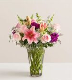 A vibrant floral arrangement in a clear glass vase featuring pink lilies, light pink roses, purple carnations, and white snapdragons accented with baby's breath and eucalyptus leaves.