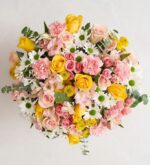 A top-down view of a lush, vibrant floral arrangement featuring yellow and pink roses, pink carnations, white daisies, and green eucalyptus leaves against a light neutral background.