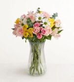 A lush floral arrangement featuring pink roses, white daisies, yellow carnations, and purple alstroemeria in a clear glass vase with a ribbed texture.