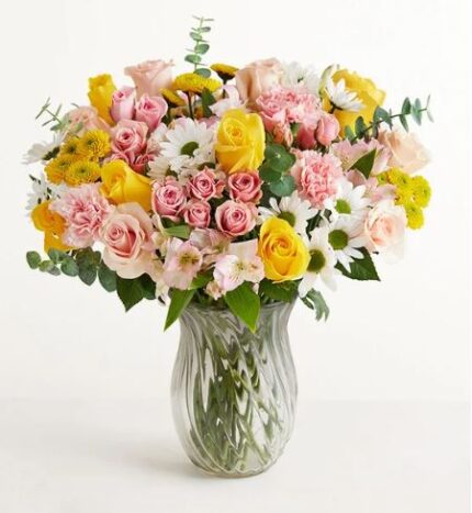 A lush and vibrant floral arrangement featuring pink roses, yellow roses, pink carnations, and white daisies in a clear swirled glass vase.