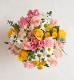 A top-down view of a vibrant floral bouquet featuring a mix of yellow roses, pink carnations, white daisies with green centers, and pink alstroemeria, accented with green eucalyptus leaves against a neutral light background.