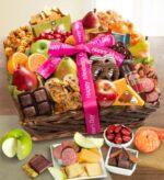 A deluxe Mother's Day gift basket overflowing with fresh fruit, gourmet sweets, and savory snacks, tied with pink "Happy Mother's Day" ribbons. Items include apples, pears, oranges, chocolate-covered pretzels, cookies, cheese, crackers, salami, caramel popcorn, and assorted chocolates in a wicker basket.