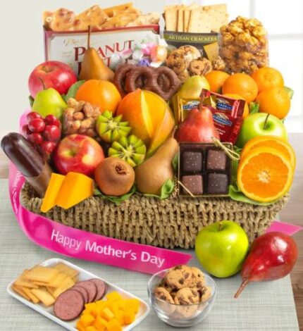 Woven gift basket filled with fresh fruit including apples, oranges, pears, kiwi, and mango, plus chocolates, cookies, crackers, and pretzels, with a pink "Happy Mother's Day" ribbon in front.