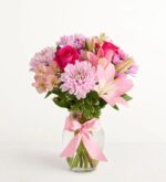 A lush bouquet of pink and purple flowers including roses, lilies, and chrysanthemums in a clear glass vase tied with a satin pink ribbon, set against a plain white background.