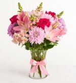 A vibrant floral arrangement featuring pink lilies, magenta roses, and light purple chrysanthemums in a clear glass vase tied with a satin pink ribbon.