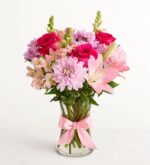 A vibrant bouquet of pink and hot pink flowers, including roses, lilies, and chrysanthemums, arranged in a clear glass vase tied with a soft pink satin bow.