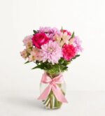 A vibrant floral arrangement featuring pink roses, carnations, and alstroemeria in a clear glass vase tied with a decorative pink satin bow.