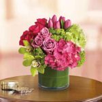 Vibrant floral arrangement in a short round green vase on a wooden table, featuring pink roses, magenta tulips, hot pink hydrangeas, lime green hydrangeas, green orchids, and small green filler flowers.