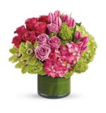A round floral arrangement in a short green glass vase featuring pink and lavender roses, pink tulips, magenta orchids, green cymbidium orchids, and light green hydrangea clusters.