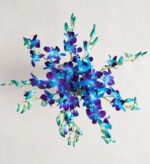 A vibrant bouquet of blue and purple Dendrobium orchids with teal-tipped petals, arranged in a starburst pattern and viewed from above against a light neutral background.