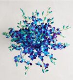 Top-down view of a lush bouquet of vibrant blue and purple dendrobium orchids with delicate green stems and leaves, arranged in a full circular burst against a soft white background.